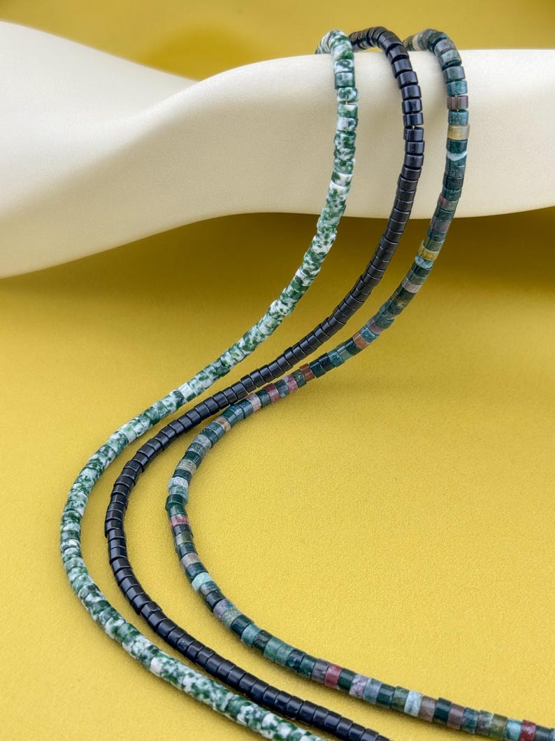 May include: Three strands of beaded necklaces featuring small cylindrical beads. One strand consists of mottled green and white beads, another of solid black beads, and the third of multicolored beads in shades of green, brown, and grey.