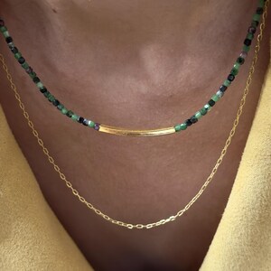 Może przedstawiać: Naszyjnik warstwowy z zawieszką w kształcie złotej belki i koralikową łańcuszkiem z zielonymi, czarnymi i fioletowymi koralikami.