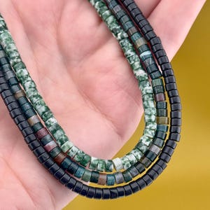 May include: A triple-strand necklace featuring small, cylindrical beads. One strand consists of mottled green and white stone beads, another features a mix of dark green, brown, and grey stone beads, and the third strand is made of solid black beads.