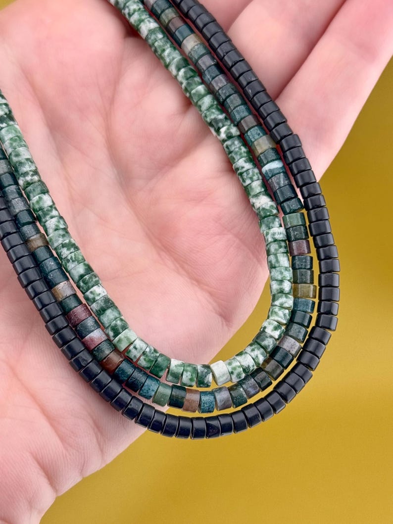 May include: A triple-strand necklace featuring small, cylindrical beads. One strand consists of mottled green and white stone beads, another features a mix of dark green, brown, and grey stone beads, and the third strand is made of solid black beads.