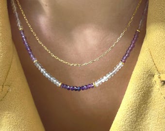 May include: A layered necklace set consisting of a delicate gold chain and a beaded strand featuring faceted purple amethyst and clear quartz beads, accented with small gold-toned spacers, resting against a yellow textured fabric garment.