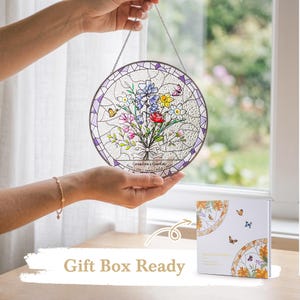 May include: A round stained glass window art piece with a floral bouquet design in shades of blue, pink, yellow, and red. The piece is held up by a chain and has a purple border. The text "Grandma's Garden" is visible. A gift box is also present.
