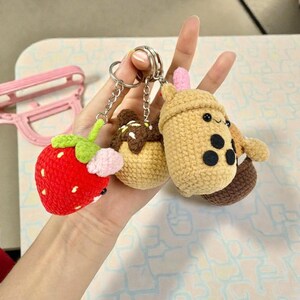 May include: A collection of handmade crochet keychains featuring a red strawberry, a chocolate-covered dessert, and a boba tea cup. The keychains are attached to silver key rings. The strawberry has green leaves and pink accents.