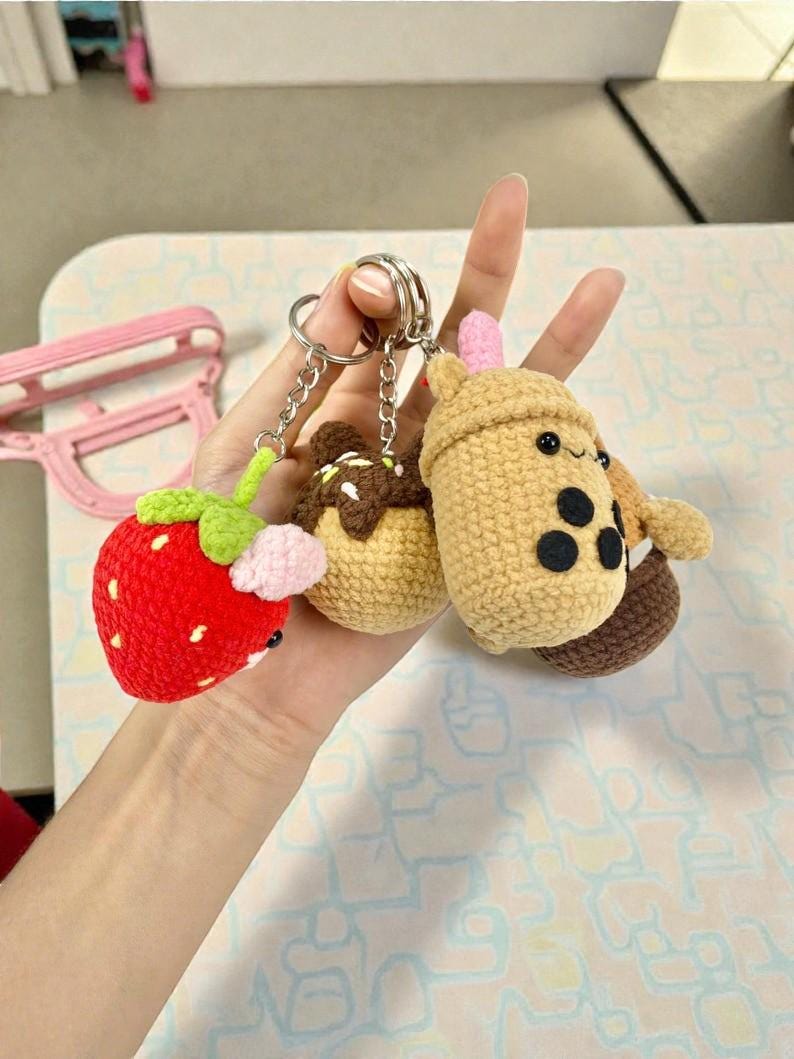 May include: A collection of handmade crochet keychains featuring a red strawberry, a chocolate-covered dessert, and a boba tea cup. The keychains are attached to silver key rings. The strawberry has green leaves and pink accents.