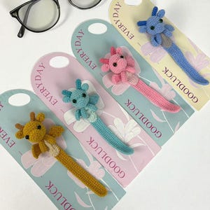 以下が含まれることがあります： 手作りの編みぐるみのしおりが4つ。それぞれウーパールーパーとキリンの形をしており、異なる配色が施されています。「GOODLUCK」と「EVERYDAY」の文字が入ったパステルカラーのカードに配置されています。黒縁のメガネが背景にあります。