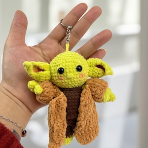 May include: A handmade crochet keychain featuring a character with large ears, black eyes, and a brown coat. The keychain is yellow and brown, with a silver keyring attached. The character's face has pink cheeks.