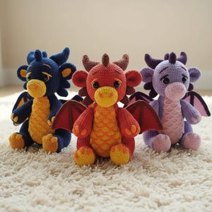 Pode incluir: Três brinquedos de dragão de crochê feitos à mão em azul, vermelho e roxo. Cada dragão tem asas, chifres e uma barriga amarela. Os dragões estão sentados em um tapete branco e fofo.