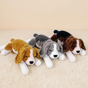 Puede incluir: Tres peluches de perros beagle en tonos marrón, gris y negro, cada uno con detalles blancos en las patas y la cara. Los perros están tumbados sobre una superficie color crema, con los ojos cerrados.