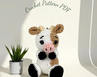 May include: A handmade crochet cow with white, brown, and black details. The cow has a pink nose and black eyes. The text "Crochet Pattern PDF" is written in cursive above the cow.