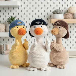May include: Three crocheted duck toys standing side by side. The left duck is yellow with a blue cap, the centre duck is white with a black cap, and the right duck is tan with a brown cap. Each duck has an orange beak and small black eyes.