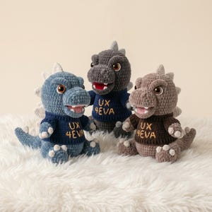May include: Three crocheted dinosaur plush toys in blue, dark gray, and light brown. Each dinosaur features a textured body, a row of dorsal spikes, and a sweater with the embroidered text UX 4EVA. The toys have brown eyes and open mouths.