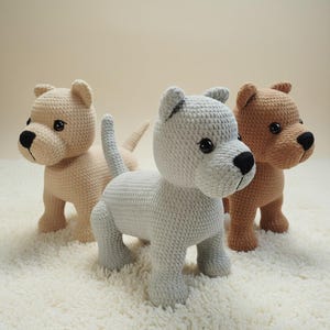 May include: Three crocheted dog figurines in beige, grey, and brown. Each dog has a black nose and button eyes. The dogs are standing on a white, textured surface. The dogs are handmade and appear to be toys.