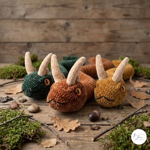 Puede incluir: Tres serpientes de peluche hechas de hilo de terciopelo suave en verde oscuro, cobre y amarillo mostaza. Cada serpiente presenta dos cuernos de color beige claro en la cabeza y ojos de color naranja brillante con pupilas negras y textura.