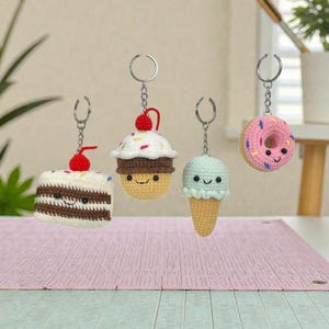 May include: Four crochet keychains shaped like desserts. The keychains include a slice of cake with a cherry, a cupcake with a cherry, an ice cream cone, and a donut. Each keychain has a silver ring and chain. The desserts have smiling faces.
