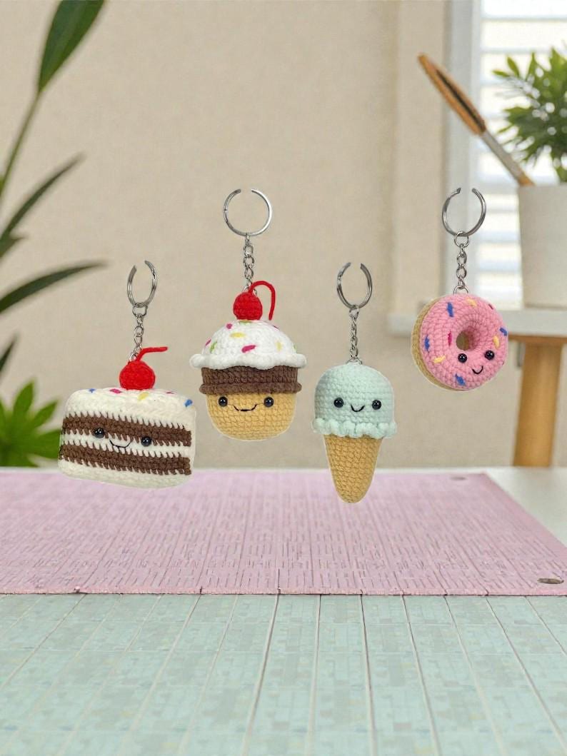 May include: Four crochet keychains shaped like desserts. The keychains include a slice of cake with a cherry, a cupcake with a cherry, an ice cream cone, and a donut. Each keychain has a silver ring and chain. The desserts have smiling faces.