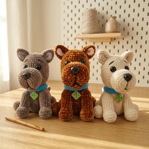 May include: Three crocheted dog plushies in gray, brown, and cream colors. Each plush toy features a blue collar with a green diamond-shaped tag, black plastic eyes, and a black nose. A wooden crochet hook lies on the surface in front of the toys.