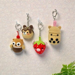 May include: Four handmade crochet keychains hang against a textured wall. The keychains are shaped like a cupcake, a takoyaki ball, a strawberry, and a bubble tea. Each keychain has a silver ring and a white hook.