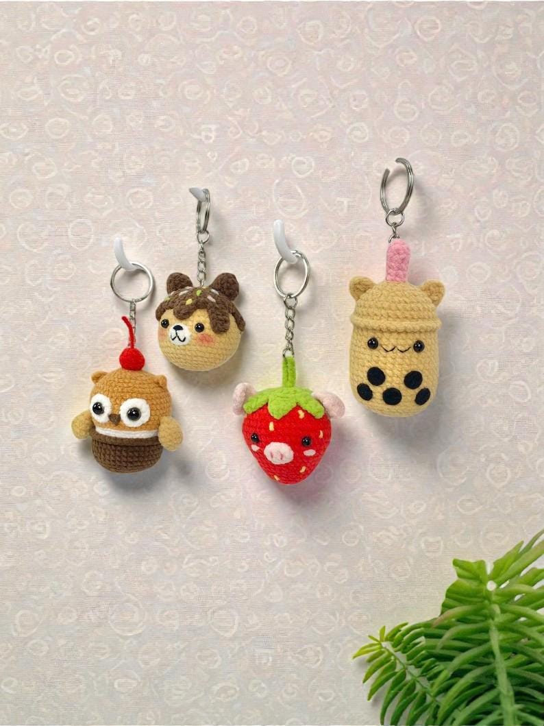 May include: Four handmade crochet keychains hang against a textured wall. The keychains are shaped like a cupcake, a takoyaki ball, a strawberry, and a bubble tea. Each keychain has a silver ring and a white hook.