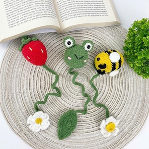 May include: Crocheted bookmarks featuring a red strawberry, a green frog, and a yellow and black bee. The bookmarks also include white flowers and a green leaf. The items are placed on a textured, circular mat.