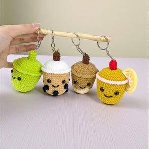 May include: A collection of four crocheted keychains, each designed to resemble a different beverage or treat. The keychains are in the shape of a green acorn, a boba tea, a coffee, and a lemon drink. Each keychain has a smiling face.