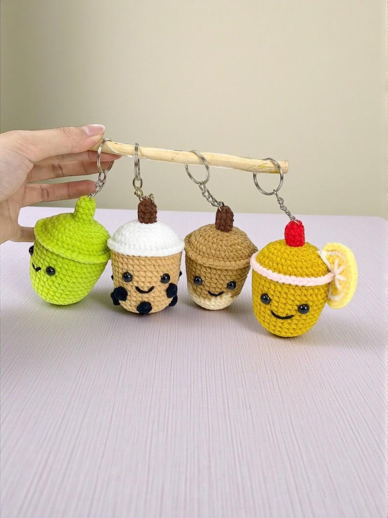 May include: A collection of four crocheted keychains, each designed to resemble a different beverage or treat. The keychains are in the shape of a green acorn, a boba tea, a coffee, and a lemon drink. Each keychain has a smiling face.