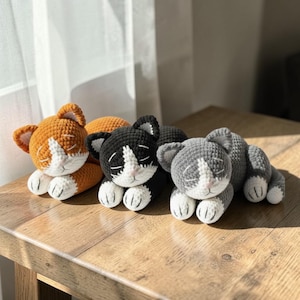 以下が含まれることがあります： オレンジ、黒、グレーの3匹のかぎ針編みの猫のおもちゃ。顔と足には白いアクセントが付いています。手作りの猫のおもちゃは目を閉じて横たわっており、眠そうな外観になっています。おもちゃは木製の表面にあります。