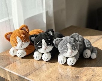 May include: Three crocheted cat toys in orange, black, and gray, each with white accents on the face and paws. The handmade cat toys are lying down with their eyes closed, giving them a sleepy appearance. The toys are on a wooden surface.