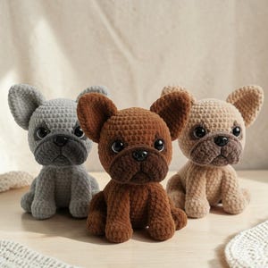 以下が含まれることがあります： グレー、ブラウン、ベージュの3色の、かぎ針編みで作られたフレンチブルドッグの置物。それぞれ大きな垂れ耳、短い鼻、黒いボタンの目があります。犬たちは前足を前に出して座っています。