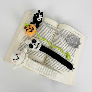 May include: Crocheted Halloween-themed bookmarks. The bookmarks feature a ghost, a skull, a jack-o'-lantern, a spider, and a spiderweb. The bookmarks are white, black, orange, and gray. The bookmarks are on top of an open book.