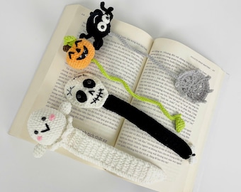 May include: A selection of crocheted Halloween-themed bookmarks. The bookmarks include a ghost, a skull, a jack-o'-lantern, a spider, and a spiderweb. The colours are white, black, orange, and grey. The bookmarks are placed on an open book.