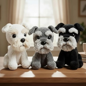 May include: Three crocheted Schnauzer dog plushies in white, grey, and black. Each has a fluffy beard, eyebrows, and a black nose. They are sitting on a wooden surface, with a window and curtains in the background.