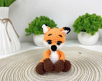 May include: A handmade crochet fox toy with orange, white, and brown yarn. The fox has black eyes and ears, and is sitting on a beige circular mat. The background includes white vases and green plants.