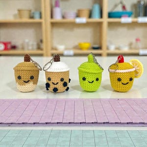 May include: Four crocheted keychains shaped like beverages. The keychains are in the shape of a boba tea, a lime drink, and a lemon drink. Each keychain has a smiling face. The boba tea keychain has black bobas. The lime drink has a green color. The lemon drink has a lemon slice.
