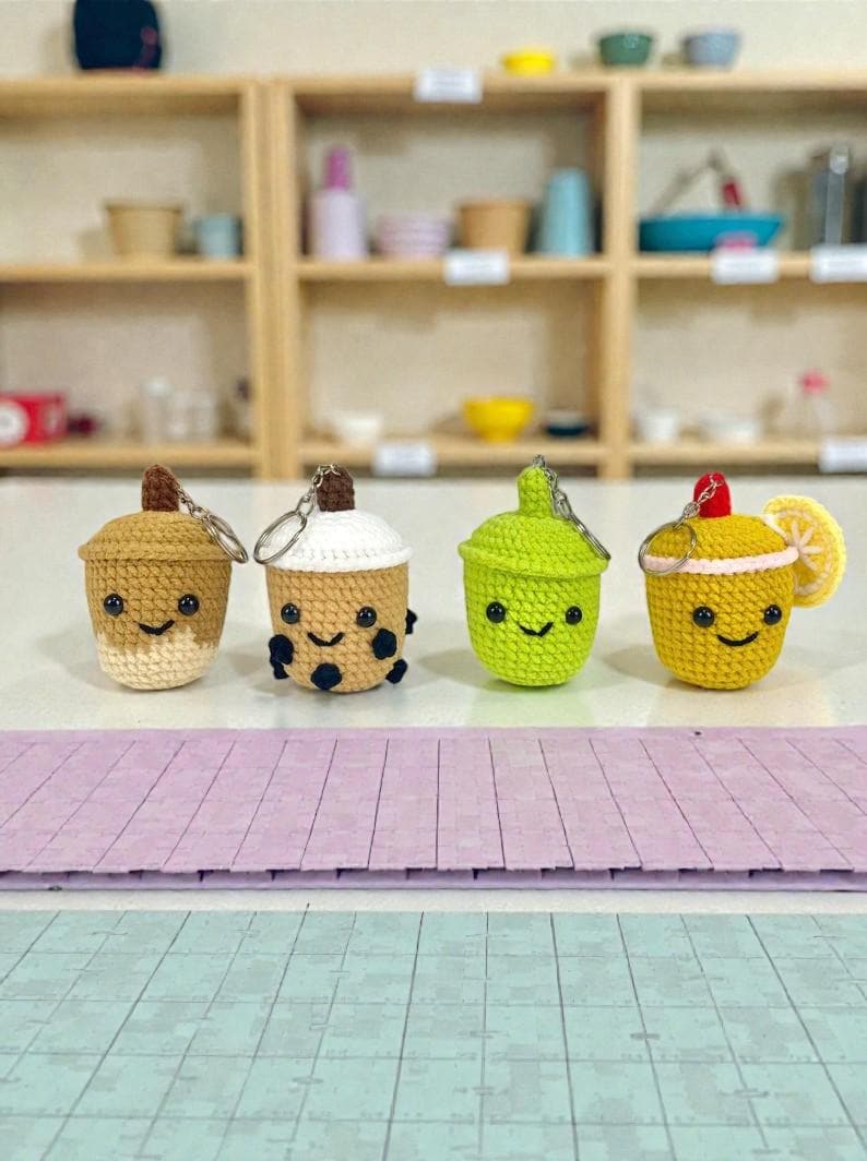 May include: Four crocheted keychains shaped like beverages. The keychains are in the shape of a boba tea, a lime drink, and a lemon drink. Each keychain has a smiling face. The boba tea keychain has black bobas. The lime drink has a green color. The lemon drink has a lemon slice.