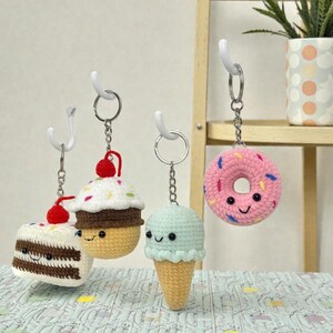 May include: Four crocheted food-themed keychains hanging from white hooks. The keychains include a slice of cake, a cupcake, an ice cream cone, and a donut. Each has a smiling face and is made with various colors of yarn. The donut is pink with colorful sprinkles.