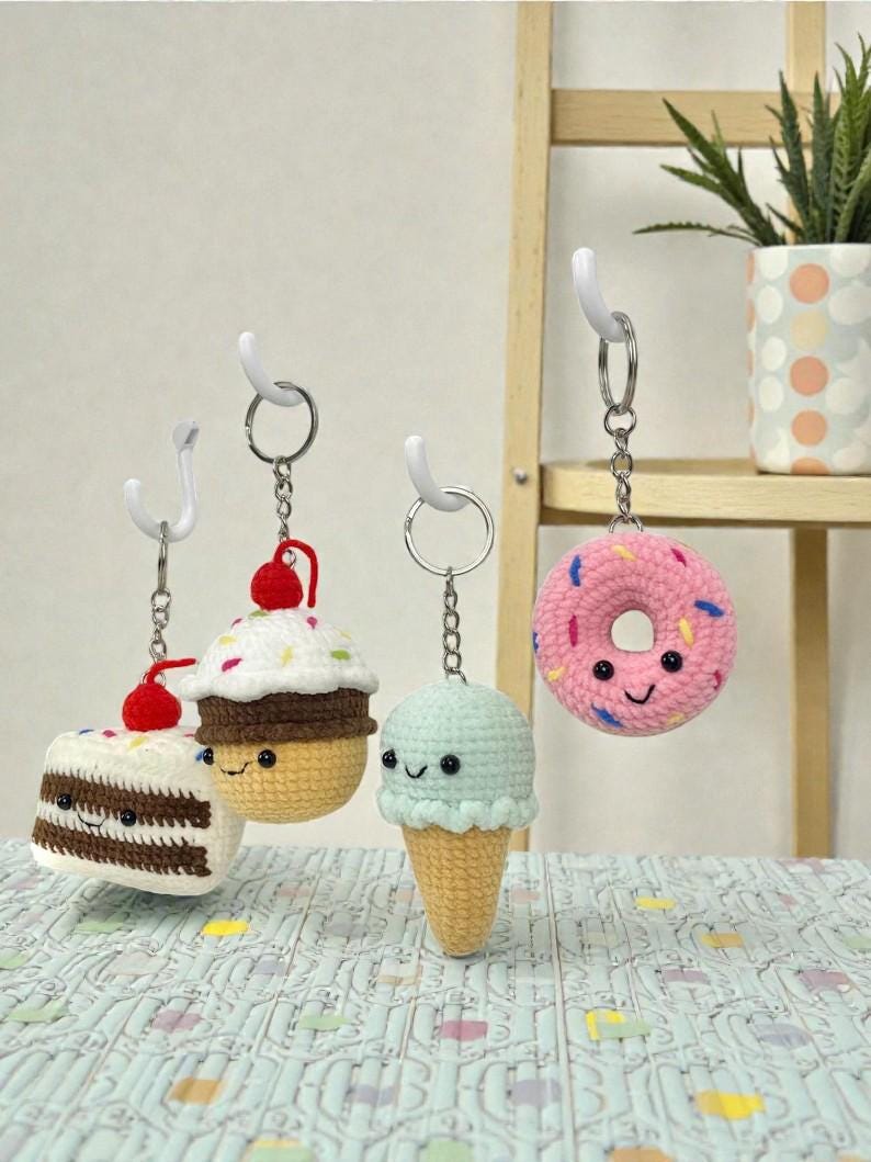 May include: Four crocheted food-themed keychains hanging from white hooks. The keychains include a slice of cake, a cupcake, an ice cream cone, and a donut. Each has a smiling face and is made with various colors of yarn. The donut is pink with colorful sprinkles.
