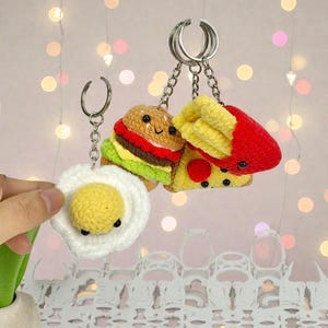 May include: Crocheted food-themed keychains, including a burger, pizza slice, fried egg, and fries. Each keychain features a silver-tone ring and chain. The burger has a brown bun, red patty, and green lettuce. The pizza has yellow cheese and red pepperoni.
