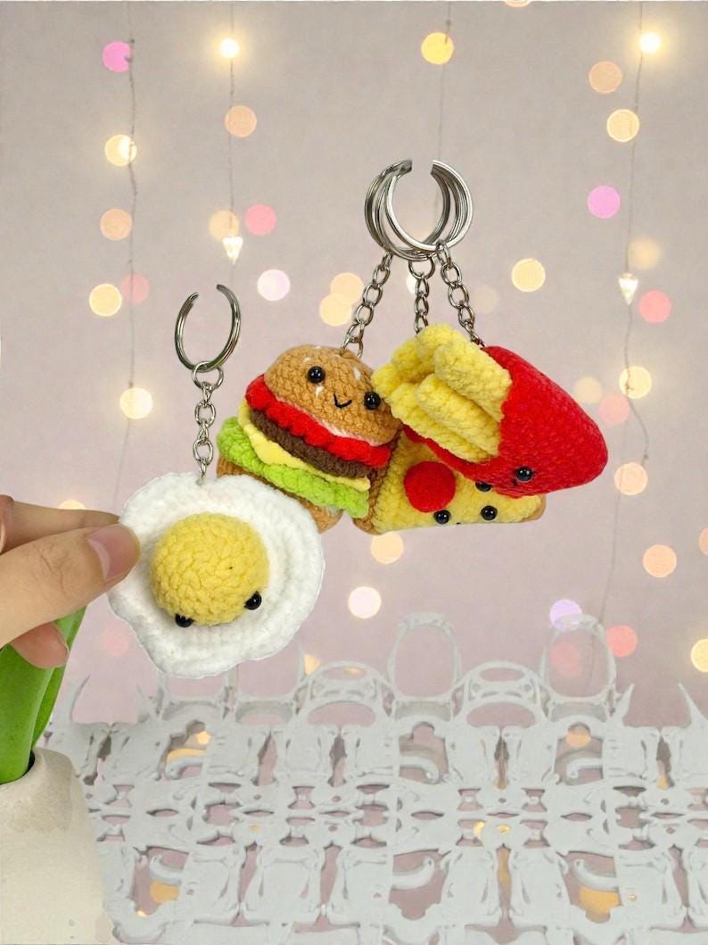 May include: Crocheted food-themed keychains, including a burger, pizza slice, fried egg, and fries. Each keychain features a silver-tone ring and chain. The burger has a brown bun, red patty, and green lettuce. The pizza has yellow cheese and red pepperoni.