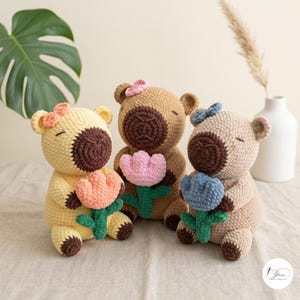 May include: Three crocheted plush capybara toys in yellow, tan, and beige, each featuring a dark brown snout, closed eyes, and a small flower accessory on the head. Each capybara holds a crocheted tulip in peach, pink, or blue with a green stem.