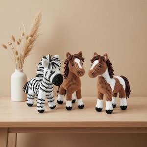 Op de afbeelding: Drie gehaakte dierenfiguren: een zebra met zwart-witte strepen en twee bruine paarden met witte markeringen. De figuren worden gepresenteerd op een lichtgekleurd houten oppervlak.