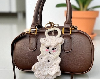 May include: A brown leather handbag with a top handle and a detachable shoulder strap. A white crochet sheep keychain is attached to the handle. The keychain has a smiling face and a flower-shaped body.