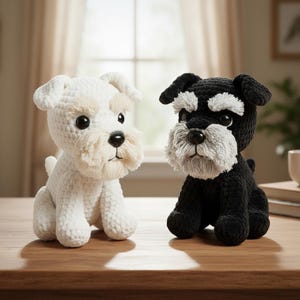Pu&ograve; includere: Due figurine di cani all'uncinetto, una bianca e una nera, sono posizionate fianco a fianco su una superficie di legno. Il cane bianco ha accenti beige, mentre il cane nero ha accenti grigi. Entrambi hanno grandi occhi scuri e barbe soffici.