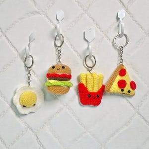 May include: Four crocheted food-themed keychains: a fried egg, a hamburger, a container of french fries, and a pizza slice. Each keychain has a metal ring and is attached to a white plastic hook. The background is a white quilted surface.