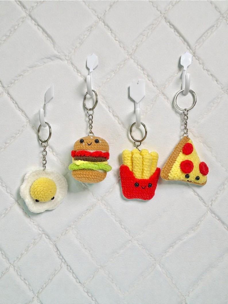 May include: Four crocheted food-themed keychains: a fried egg, a hamburger, a container of french fries, and a pizza slice. Each keychain has a metal ring and is attached to a white plastic hook. The background is a white quilted surface.