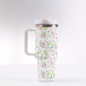 Puede incluir: Un termo blanco con asa, con un estampado floral con flores rosas, moradas y azules, hojas verdes e ilustraciones de un conejo. El termo tiene una tapa blanca y una pajita.