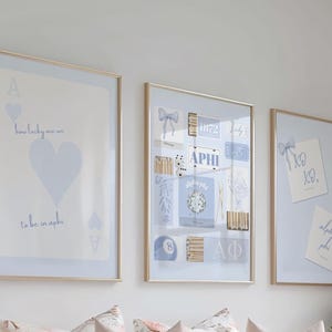 Op de afbeelding: Drie ingelijste kunstdrukken met gouden frames. De prints bevatten lichtblauwe en witte ontwerpen, waaronder speelkaartelementen, tekst en decoratieve motieven. Het algemene thema is een studentenvereniging of een college-esthetiek.