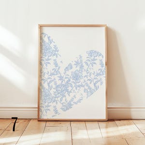 May include: Framed art print featuring a heart-shaped design filled with light blue floral patterns on a cream background. The artwork is encased in a natural wood frame, adding a touch of elegance to the piece. The print is displayed against a white wall.