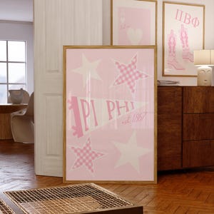 May include: Pink framed wall art with white stars and a pennant that reads "IPI PHI est 1867". Two additional framed prints are visible, one with a heart and the other with pink cowboy boots. The art is displayed in a room with wood flooring.