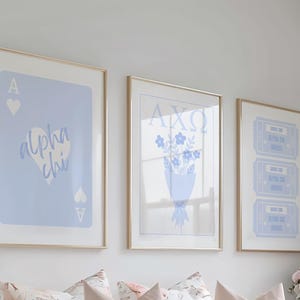 Può includere: Tre stampe artistiche incorniciate con cornici dorate. Le stampe presentano disegni azzurri su sfondo bianco. Una stampa mostra una carta da gioco con il testo "Alpha Chi". Un'altra ha un bouquet di fiori. La terza ha tre biglietti.