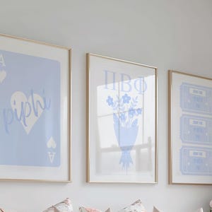 May include: Three framed art prints with gold frames. The prints feature light blue designs on a white background. One print has a playing card design with the text "piphi". Another has a bouquet of flowers. The third has three tickets.